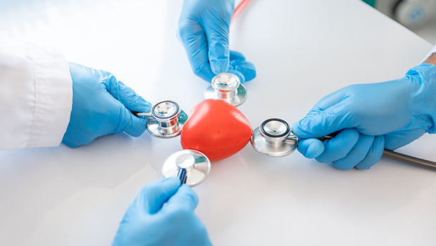 Drei H&auml;nde mit blauen Handschuhen halten jeweils ein Stethoskop an ein rotes Herz in ihrer Mitte.